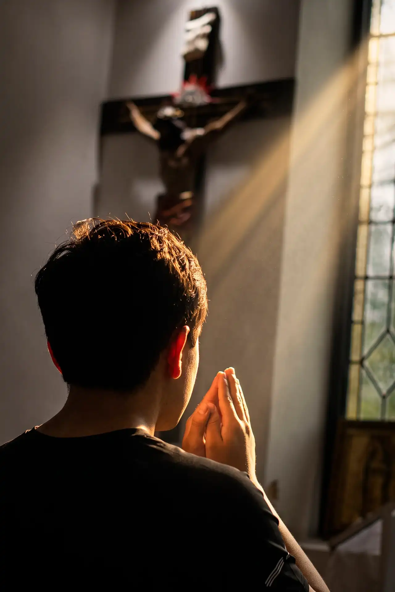 Una persona, de espaldas, junta las manos frente a un crucifijo mientras entra luz por la ventana